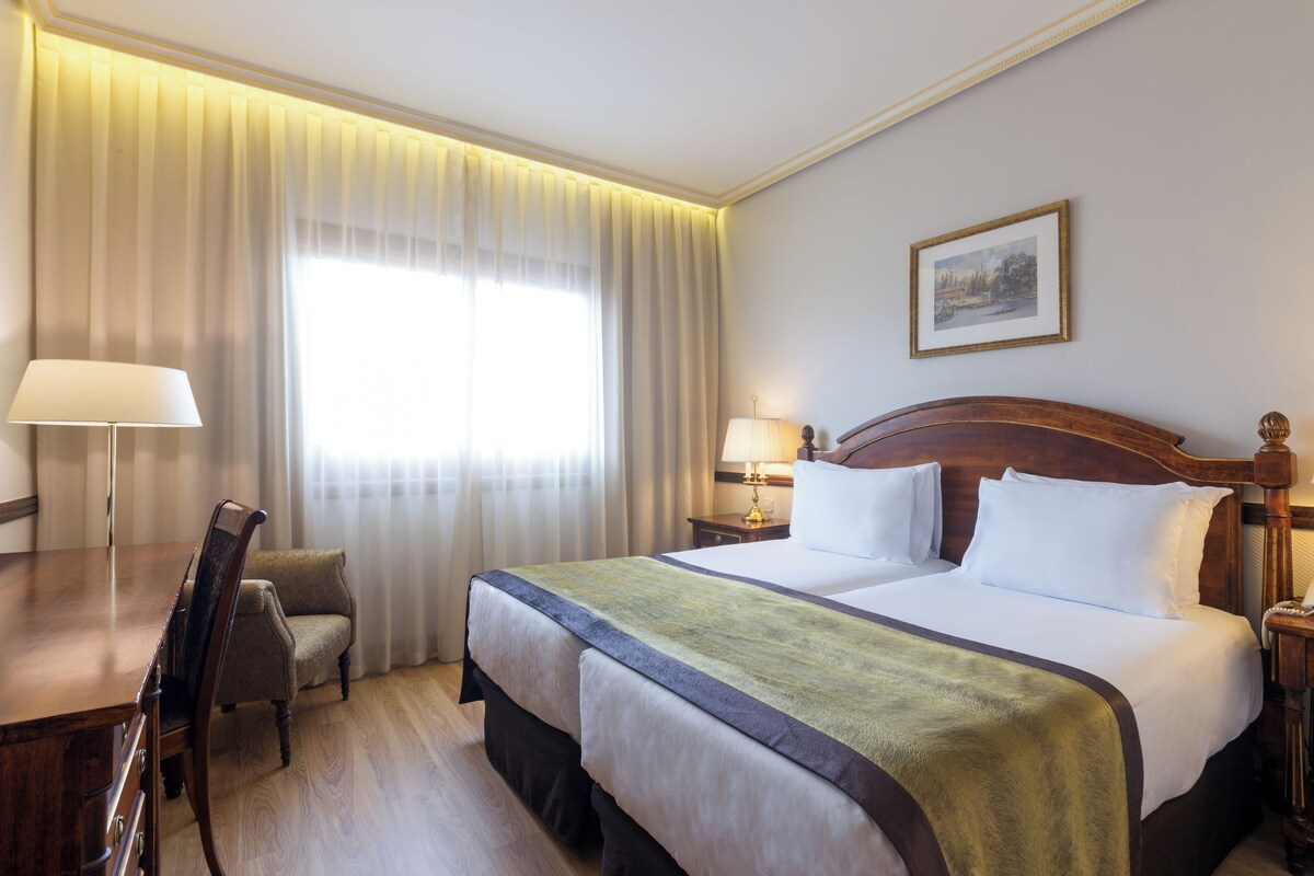 A hotel room with a large bed, a desk, a chair, and a lamp.