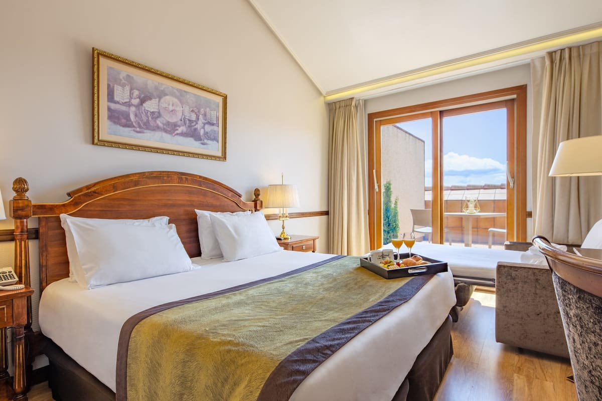 Ein Hotelzimmer mit einem großen Bett, einem Nachttisch mit Lampe, einem kleinen Tisch mit Obstkorb und Getränken sowie einem Blick ins Freie durch ein großes Fenster.