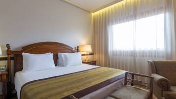A hotel room with a large bed, a wooden headboard, a bedside table with a lamp, a chair, and a window with curtains.