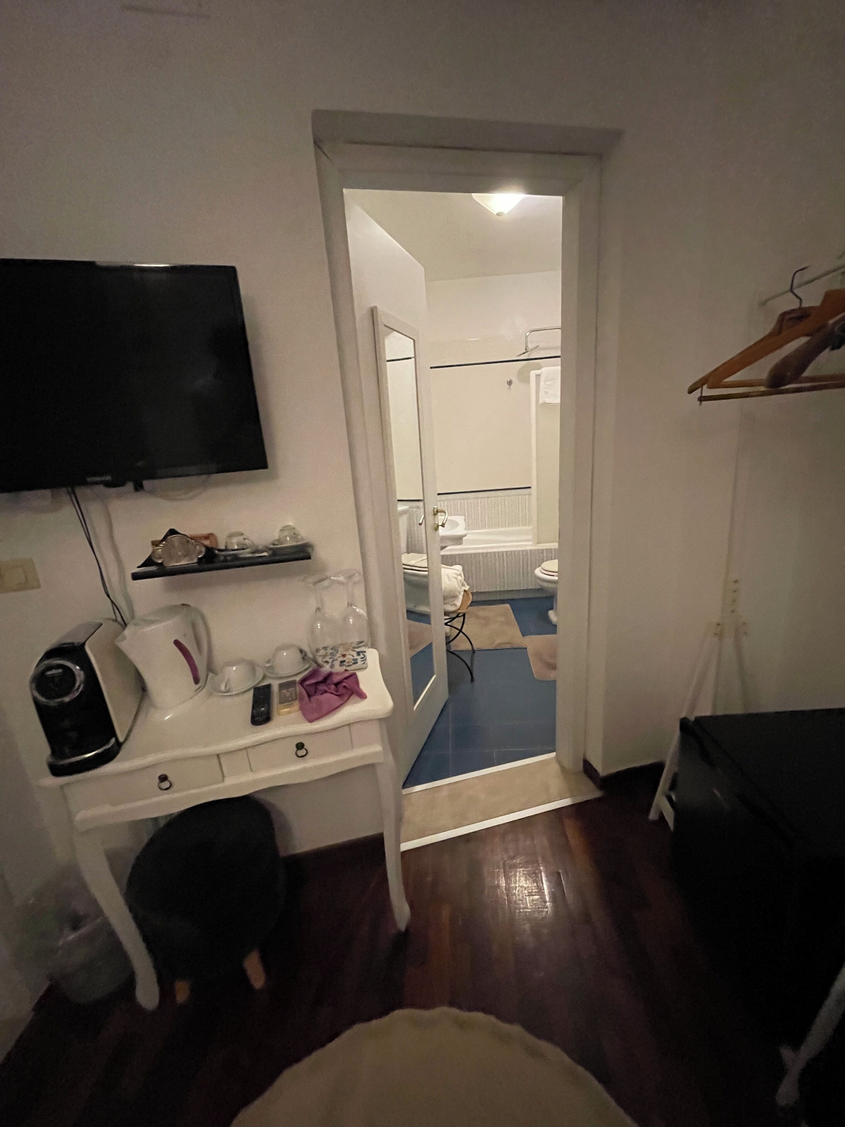 A hotel room with a TV mounted on the wall, a desk with a coffee maker, a chair, and a bed. There is a door leading to a bathroom with a mirror and a shower.