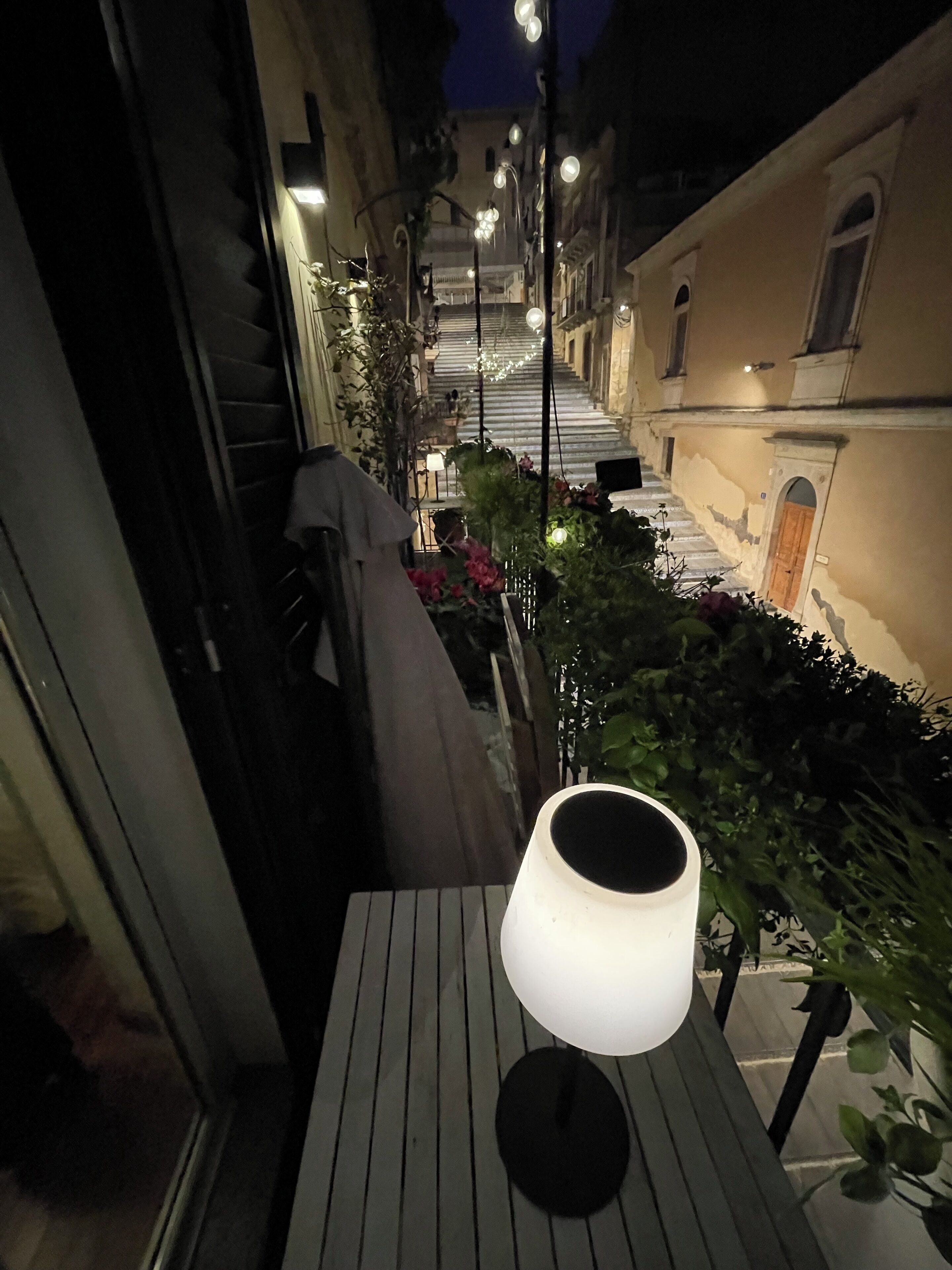 Un étroit couloir extérieur, agrémenté d’une table et d’une lampe, bordé de bâtiments et de plantes.