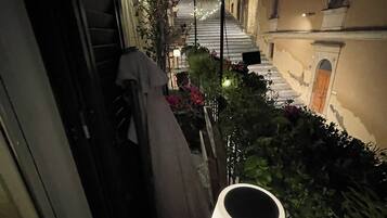 A narrow outdoor corridor with a table and a lamp, flanked by buildings and plants.