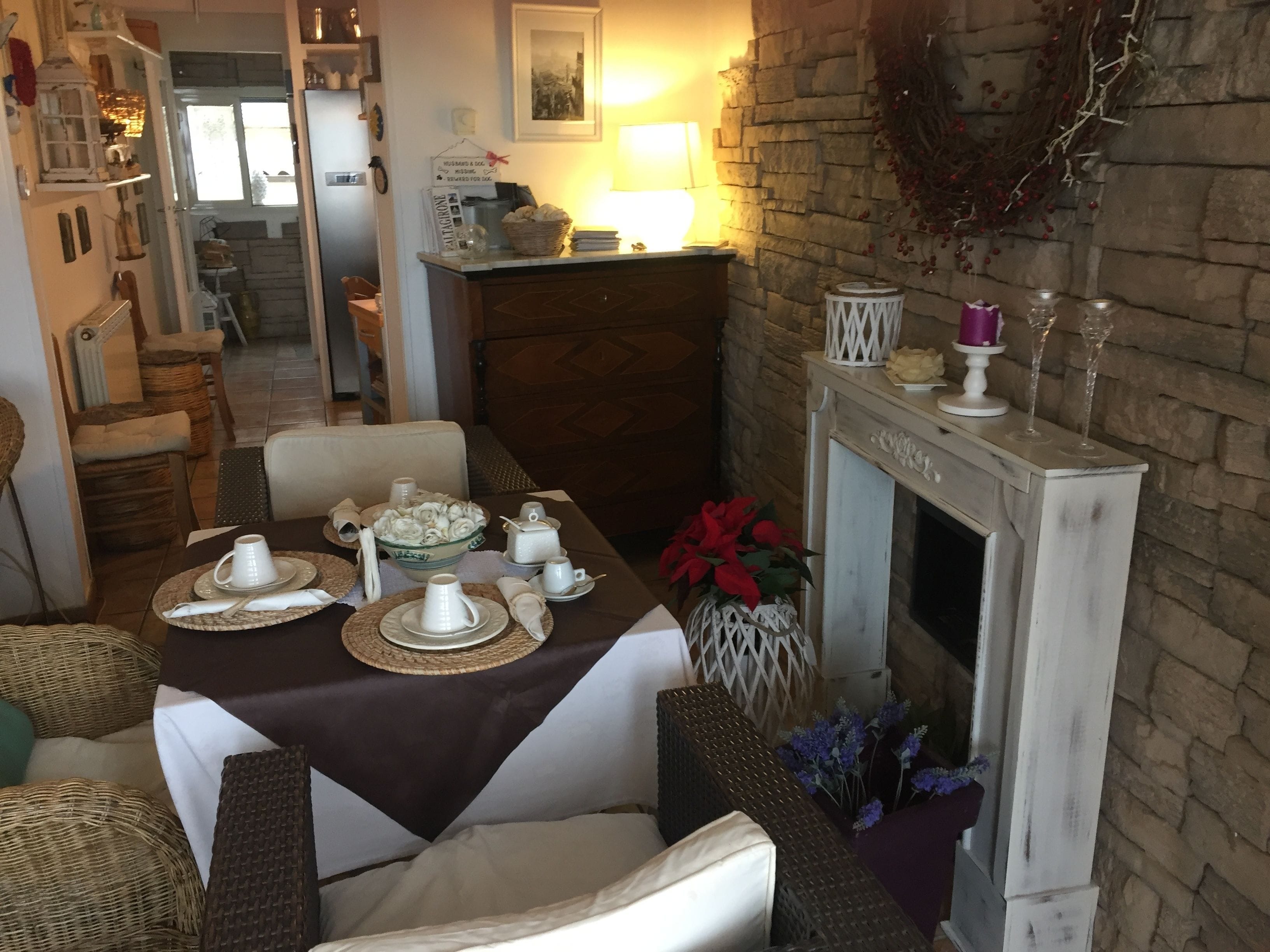 Un coin repas avec une table dressée pour quatre personnes, une cheminée, des chaises en osier et une vue sur la cuisine.