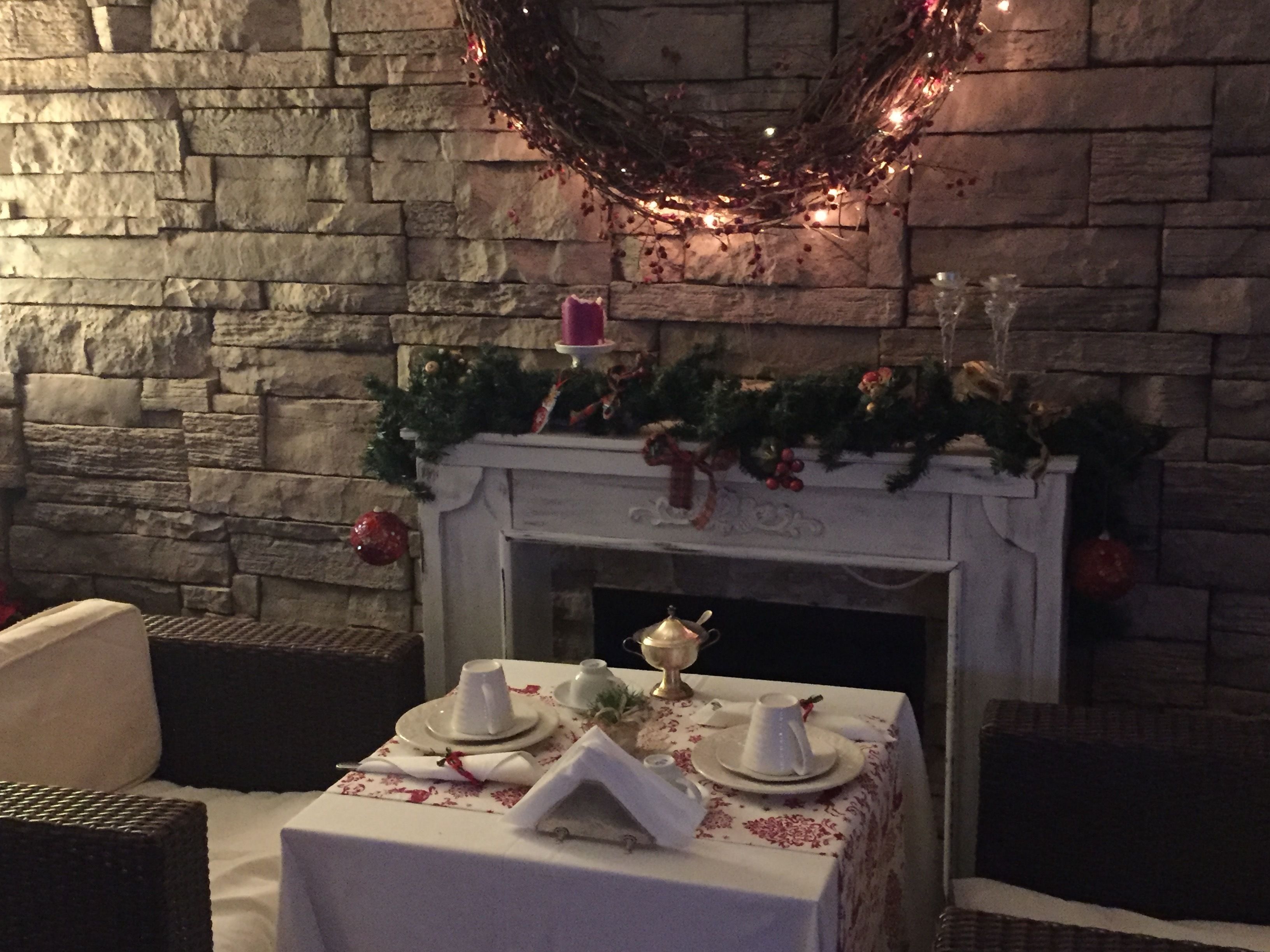 A dining area with a stone wall, a fireplace decorated with greenery and ornaments, and a table set for two with white tablecloths and plates.