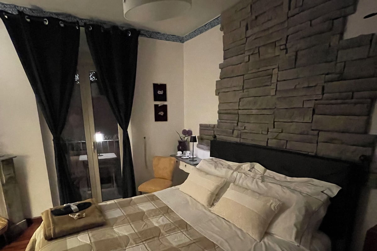 Quarto com cama, mesa de cabeceira, abajur, parede de pedra e vista pela janela com cortinas.