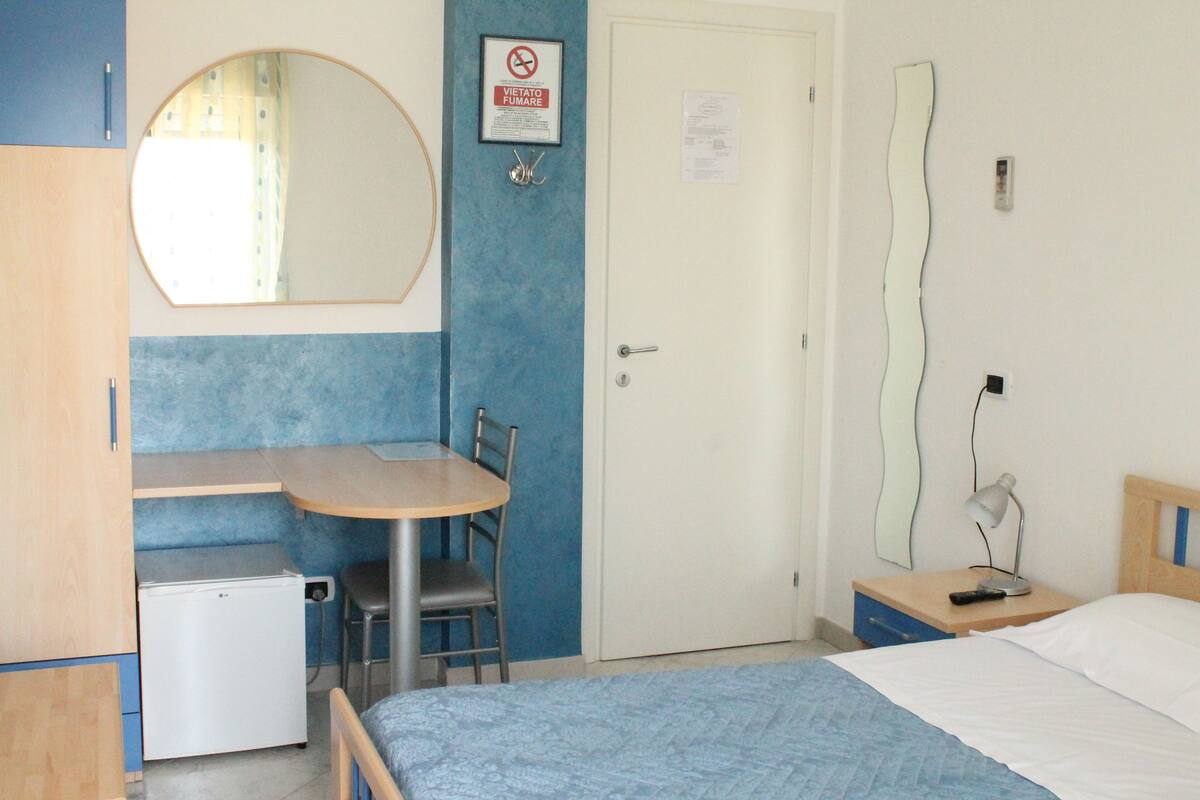 A bedroom with a bed, a desk with a chair, a small refrigerator, a door with a sign, and a wall-mounted mirror.