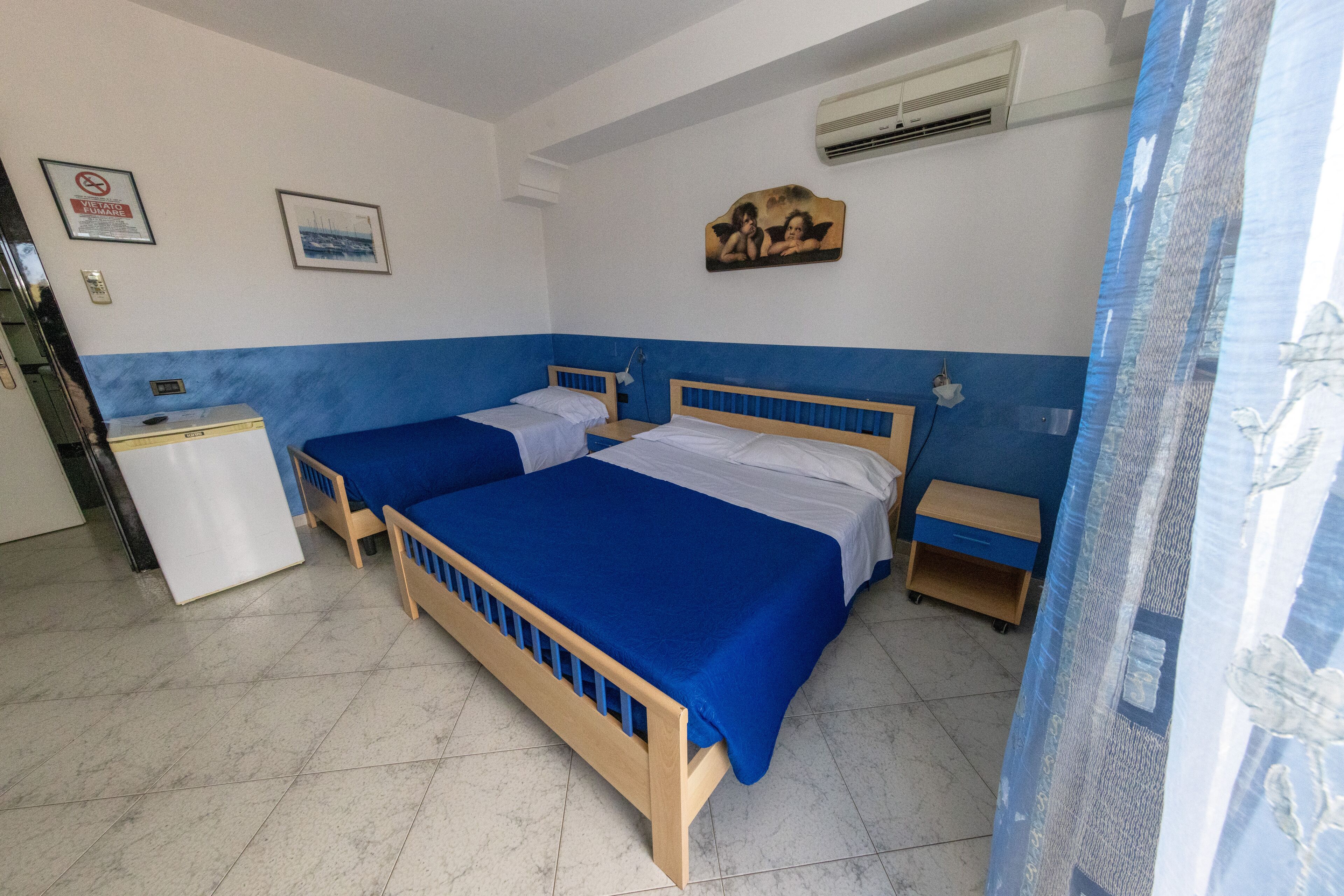 A hotel room with two beds, a small desk, a nightstand, an air conditioner, and a framed picture on the wall.