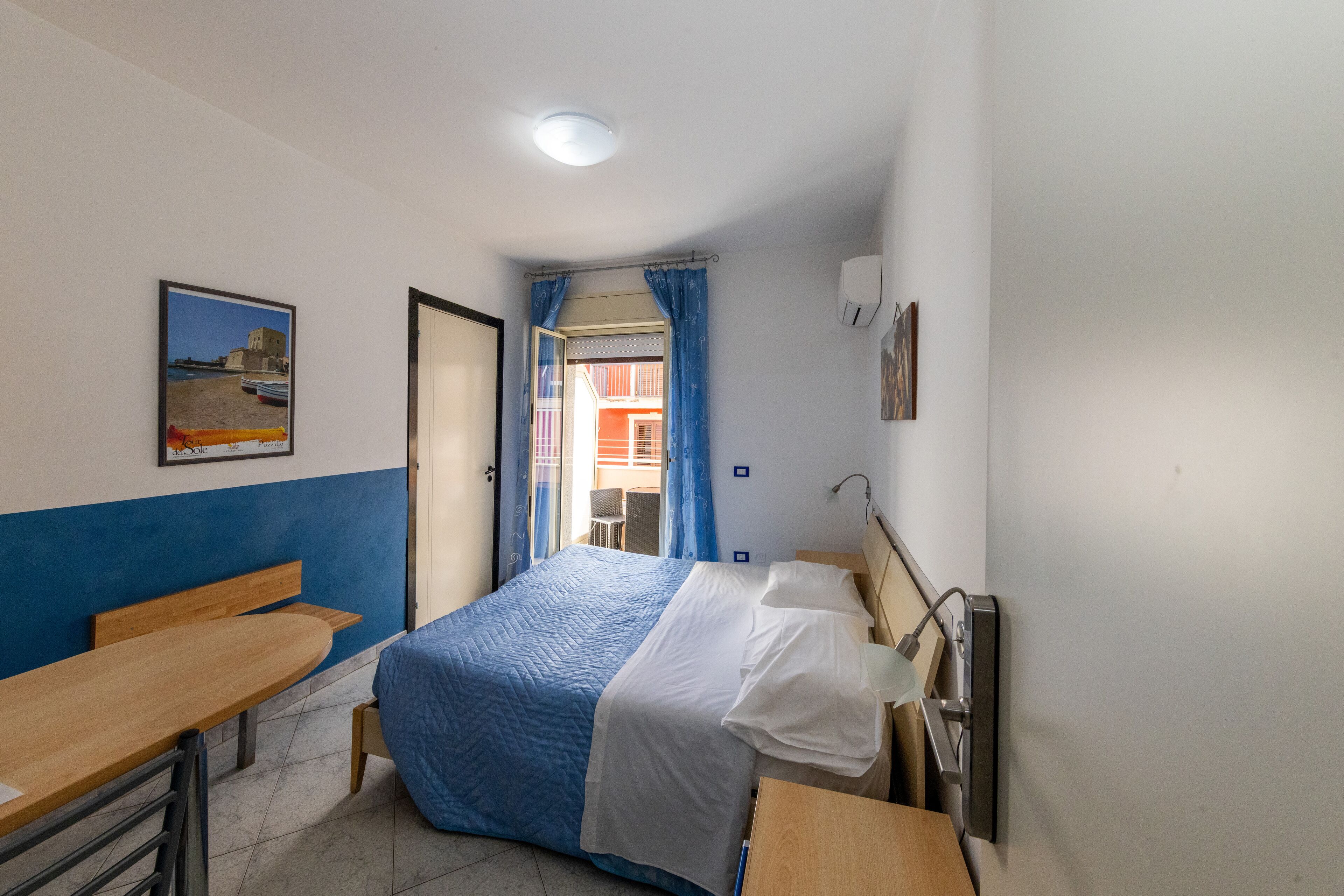 Un dormitorio con cama, escritorio y silla. Se ve una ventana con cortinas, un cuadro en la pared y una puerta que da a otra habitación.
