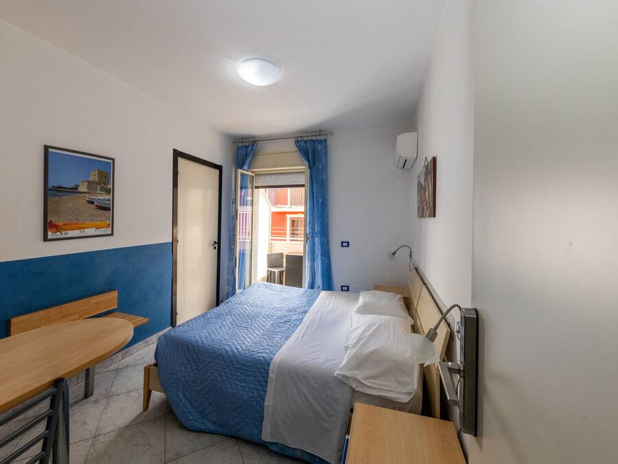Un dormitorio con cama, escritorio y silla. Se ve una ventana con cortinas, un cuadro en la pared y una puerta que da a otra habitación.