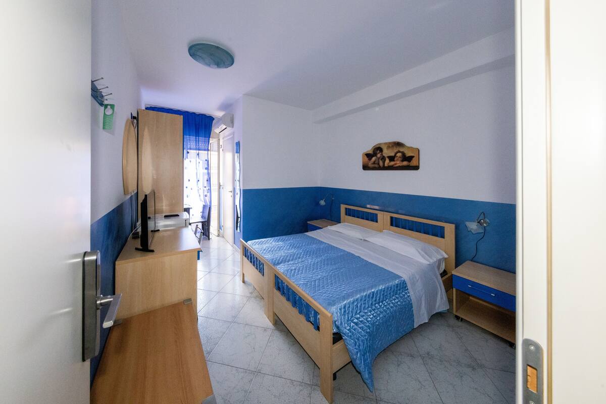 A bedroom with a wooden bed, a desk, and a chair. There is a window with blue curtains and a picture on the wall.