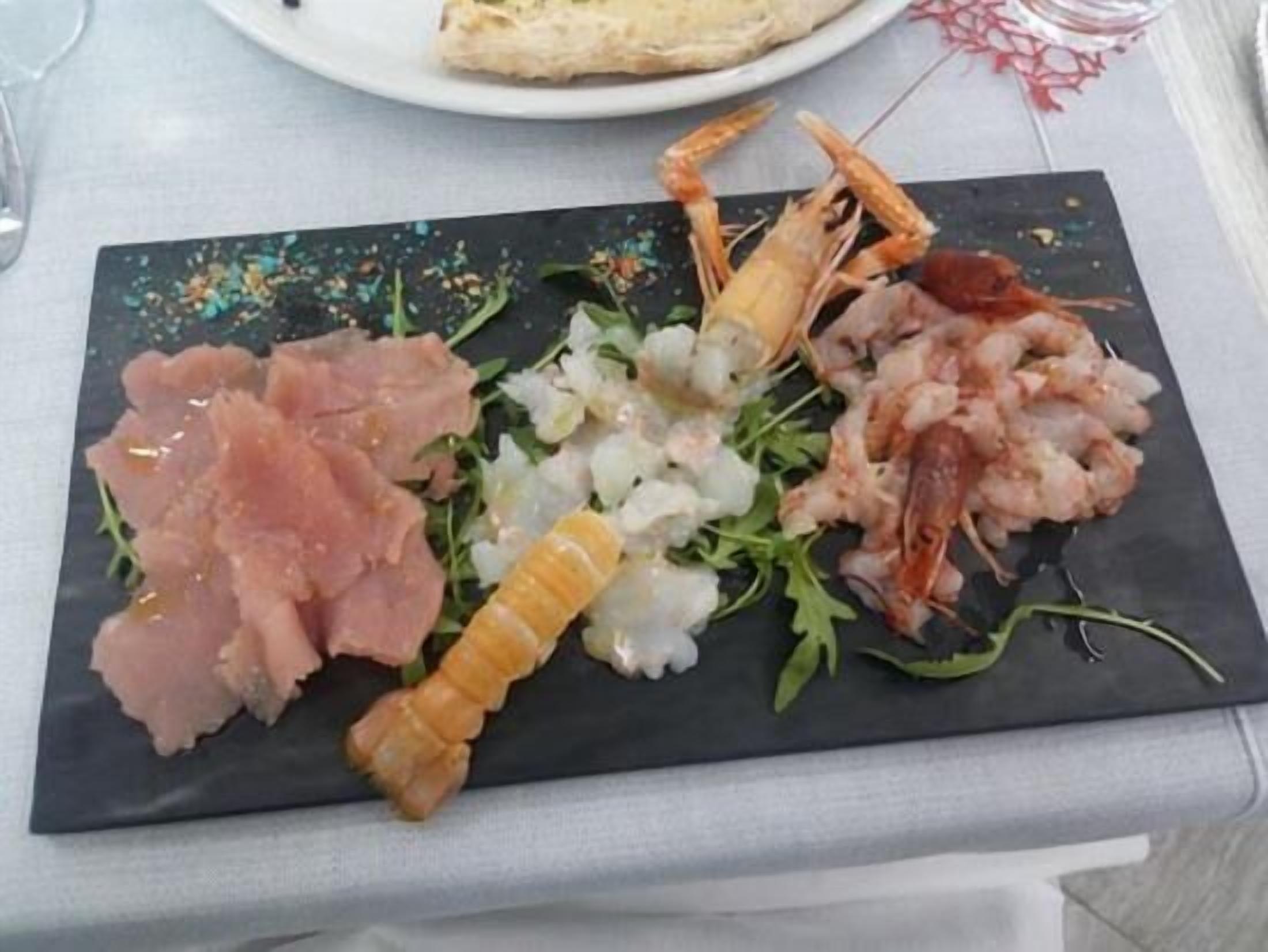 Una pizarra de pizarra negra con diversos mariscos, como camarones, vieiras y posiblemente algún tipo de carne curada, sobre un mantel blanco.