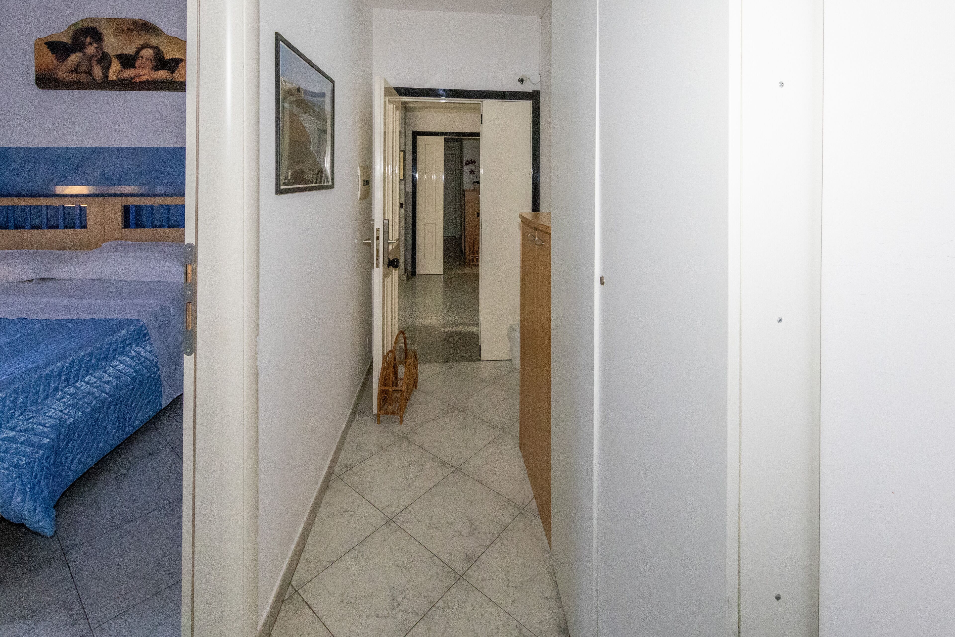Un couloir étroit, avec un lit sur la gauche, une armoire en bois sur la droite et une porte au fond qui donne sur une autre pièce.
