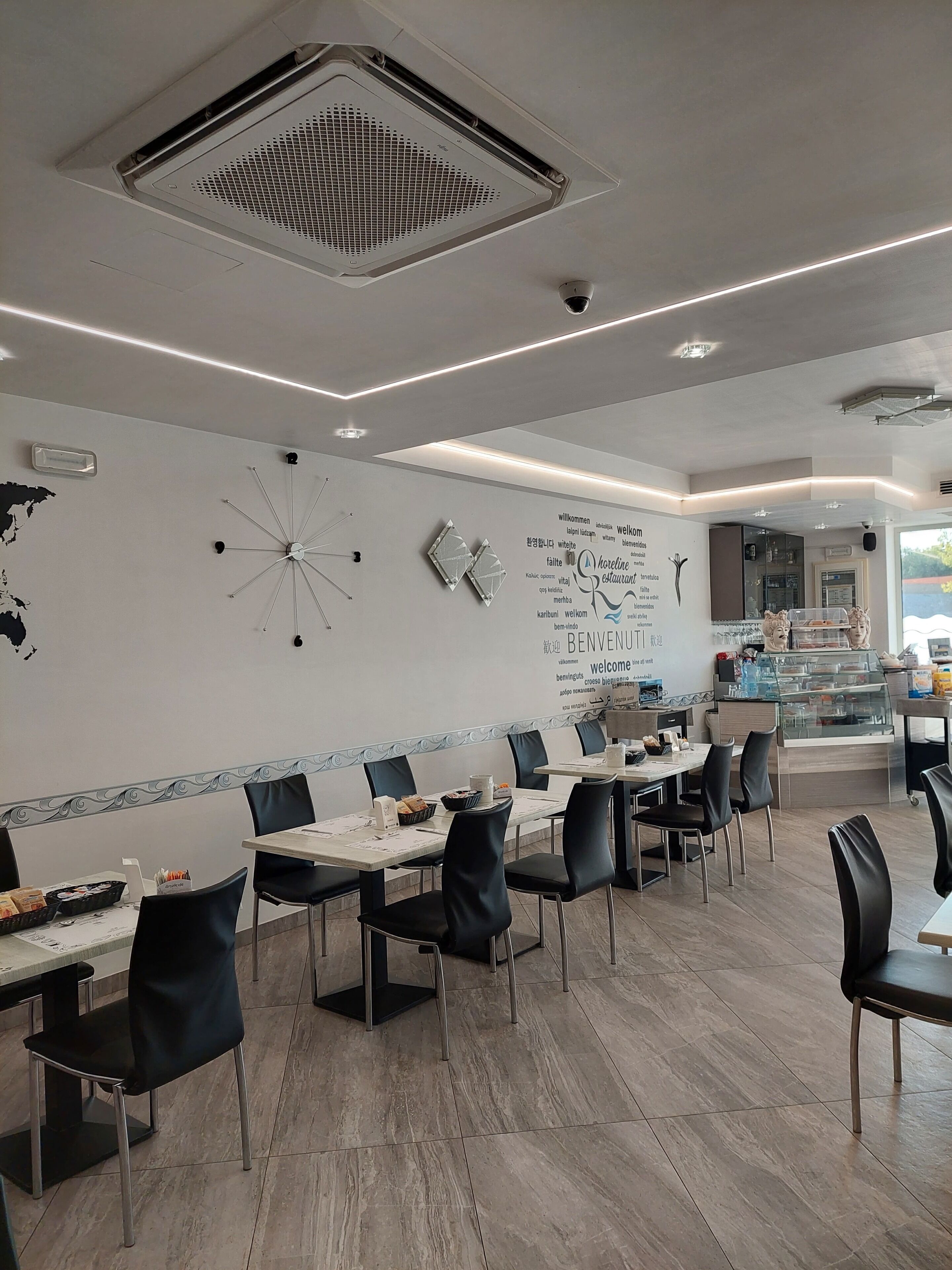 Un interior de restaurante moderno con largas mesas comunitarias, sillas negras, un mapa mundial en la pared y un tablero de menú con la palabra 'bienvenido' en varios idiomas.