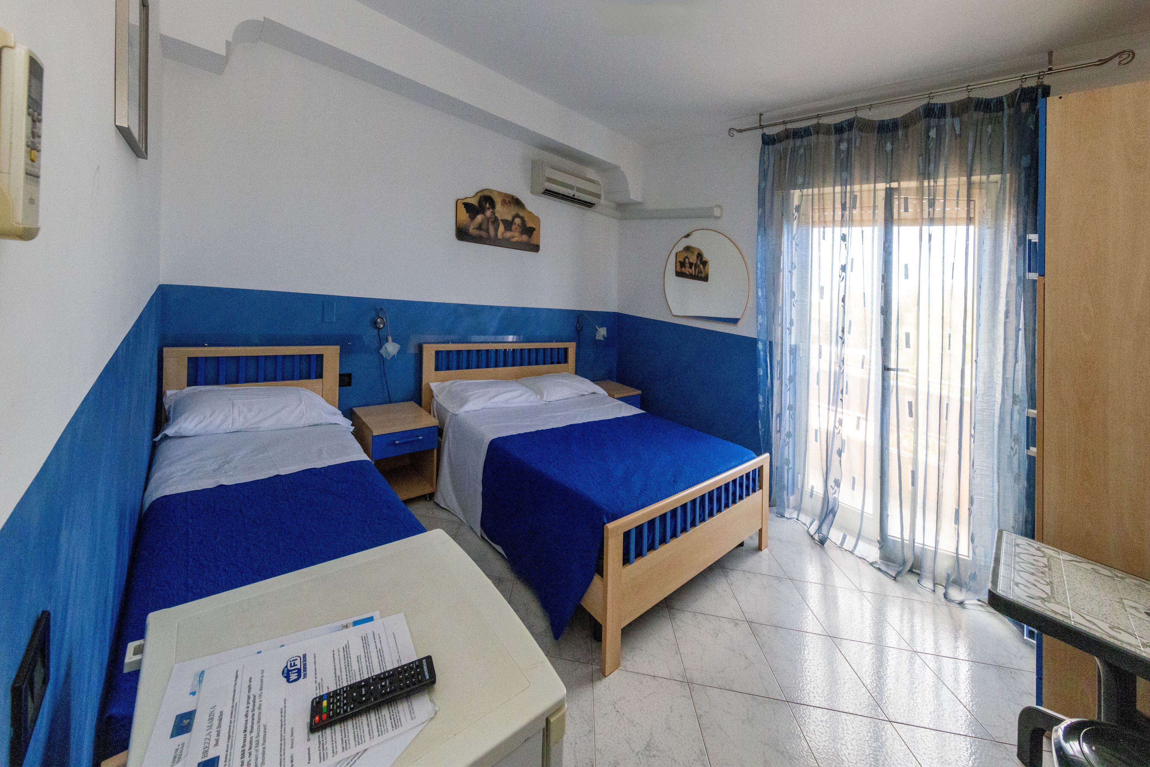 A room with two beds, a desk, and a chair. There is a window with curtains, a wall-mounted air conditioner, and a small picture on the wall.