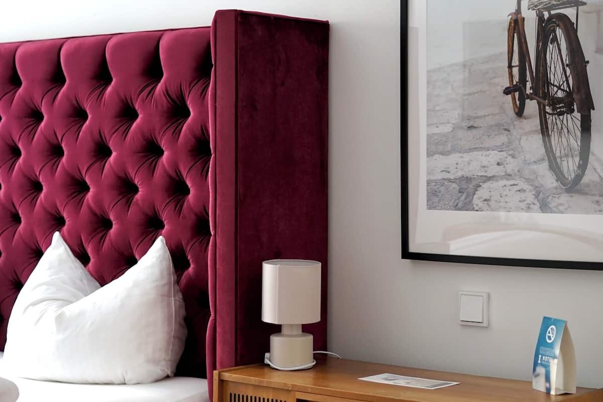 A room with a tufted headboard, a wooden nightstand with a radio, a framed picture on the wall, and a white pillow on the bed.
