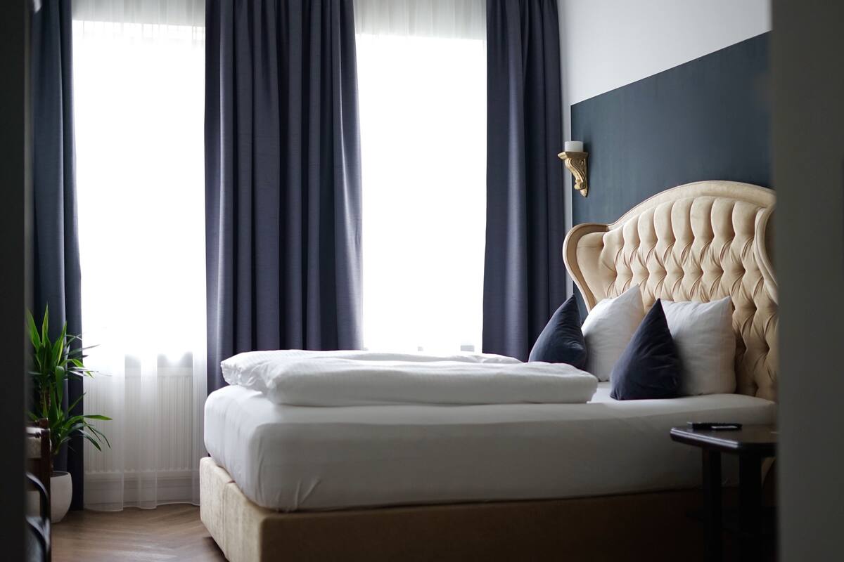 A bedroom with a large bed, dark curtains, a wall-mounted light fixture, and a potted plant.