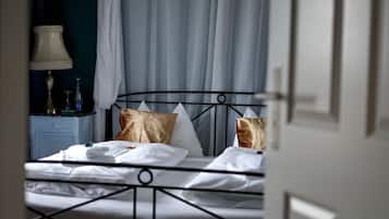 A neatly made bed with white linens and two decorative pillows, a bedside table with a lamp and a bottle, and a view of a blue curtain and a door.