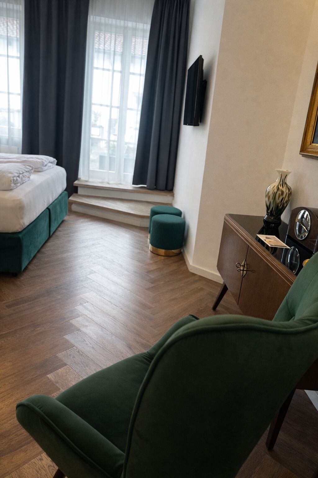 A room with a bed, a green armchair, a wooden cabinet, and a window with curtains.