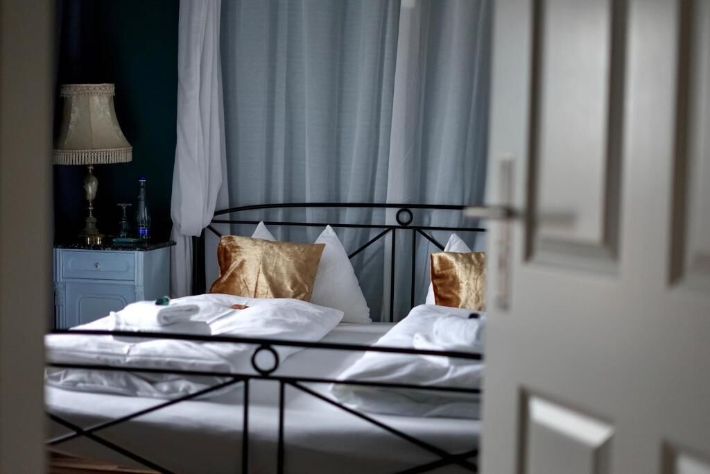 A neatly made bed with white linens and two decorative pillows, a bedside table with a lamp and a bottle, and a window with sheer curtains.