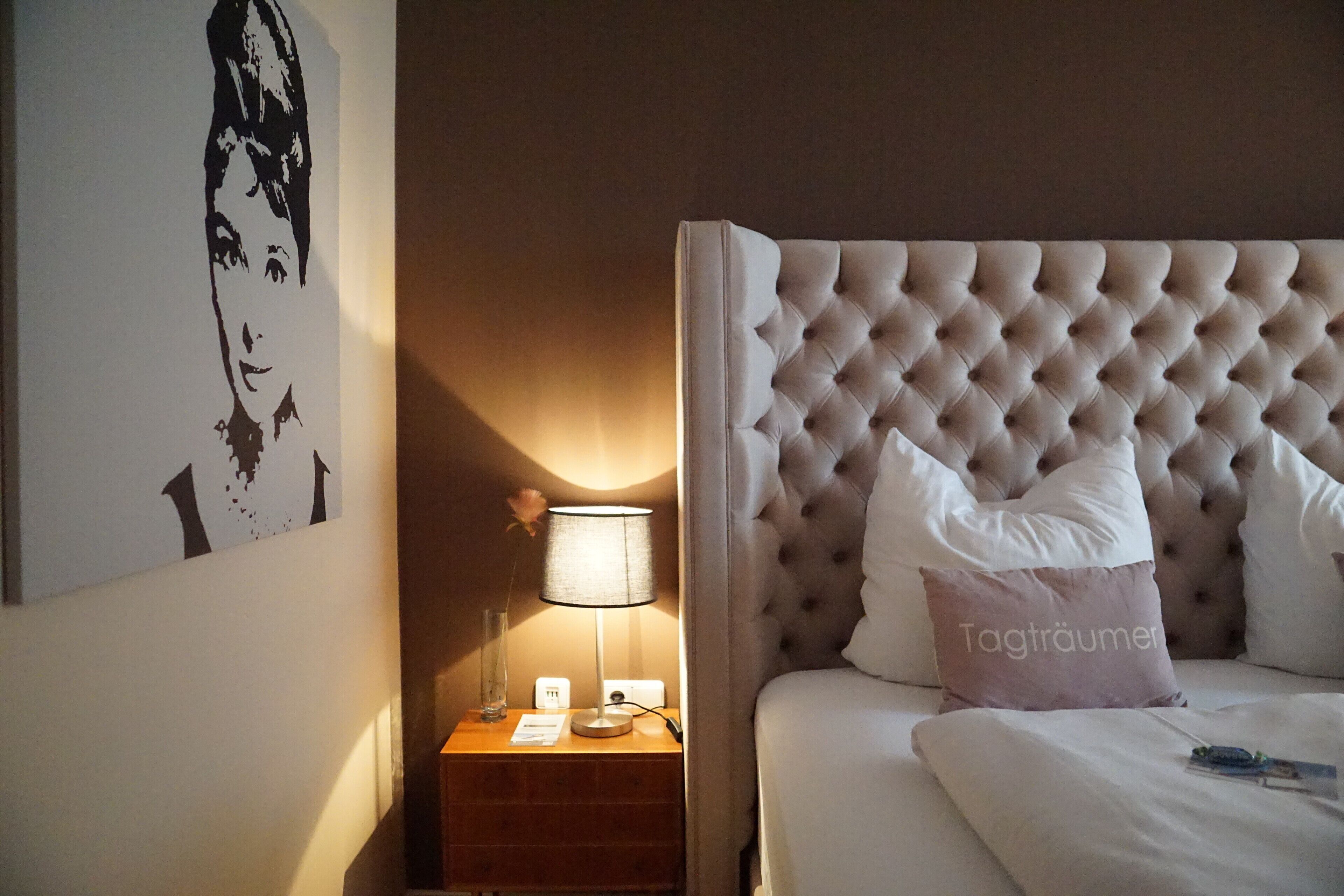 Una habitación de hotel con cabecera capitonada, una lámpara de noche, una mesita de noche y una obra de arte colgada en la pared.