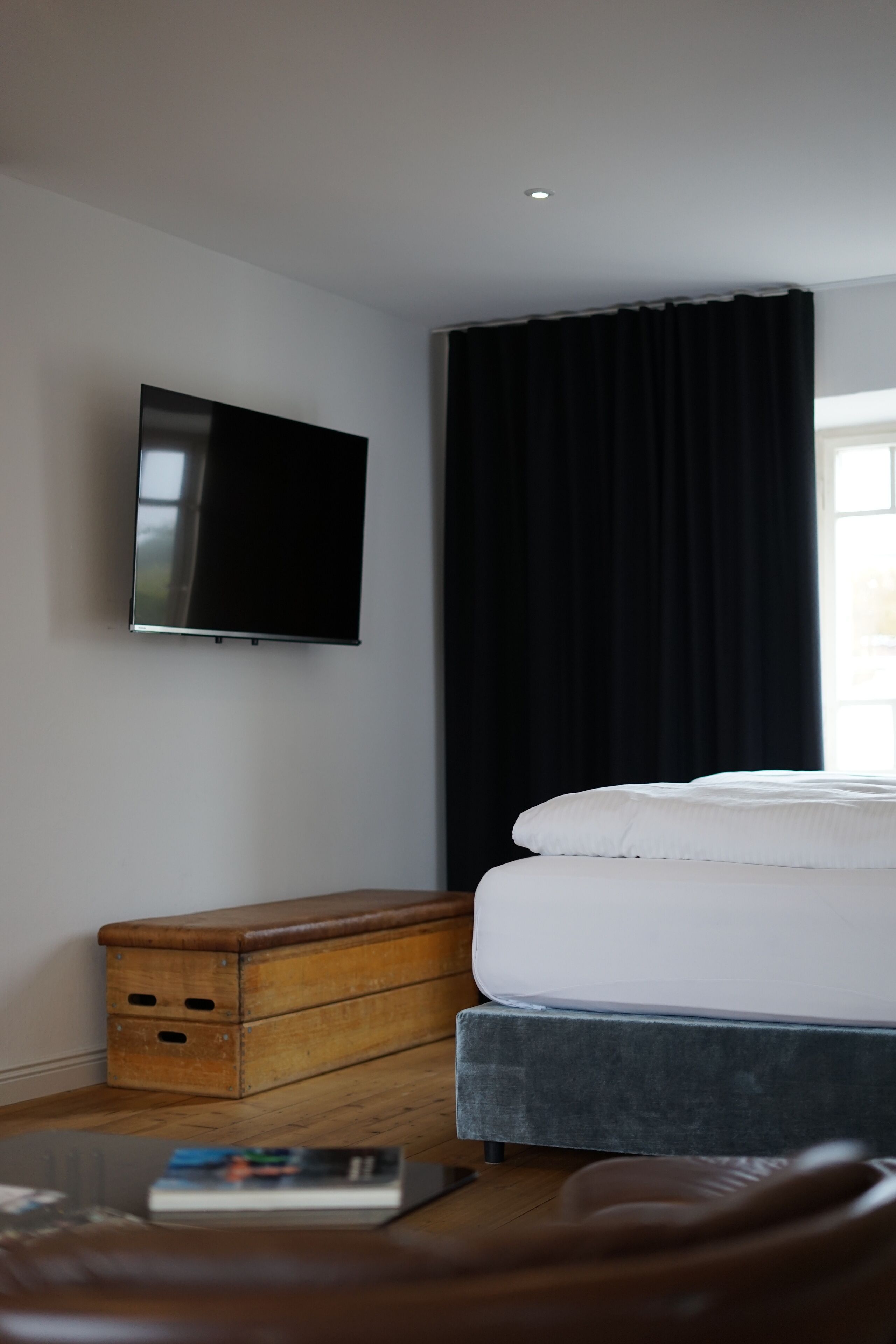 A hotel room with a bed, a flat-screen TV mounted on the wall, a wooden chest of drawers, and a window with black curtains.