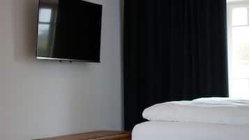 A hotel room with a bed, a flat-screen TV mounted on the wall, a wooden chest of drawers, and a window with black curtains.