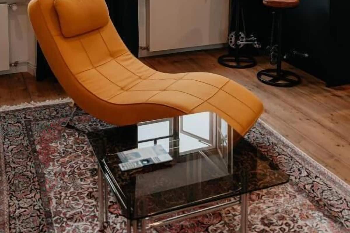A modern living room with a yellow armchair, a glass coffee table, a patterned rug, a bar area with stools, and a view of buildings through large windows.