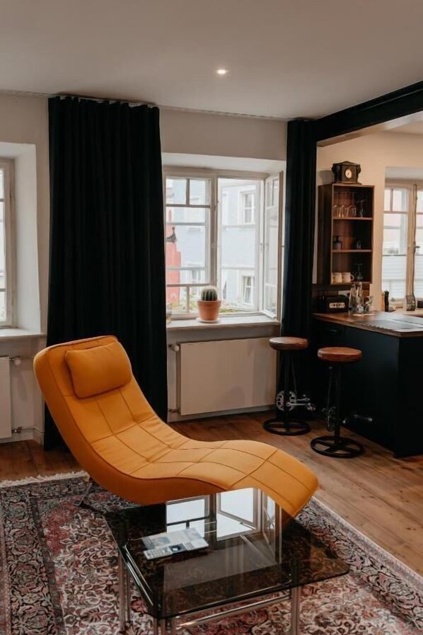 A modern living room with a yellow armchair, a glass coffee table, a patterned rug, a bar area with stools, and a view of buildings through large windows. - Hotel Mühldorf (Muhldorf am Inn)