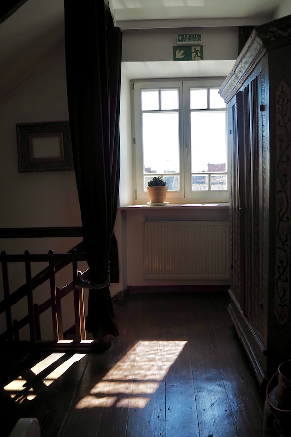 A room with a wooden floor, a window with a view, a radiator, and a door with a sign above it. - Hotel Mühldorf (Muhldorf am Inn)