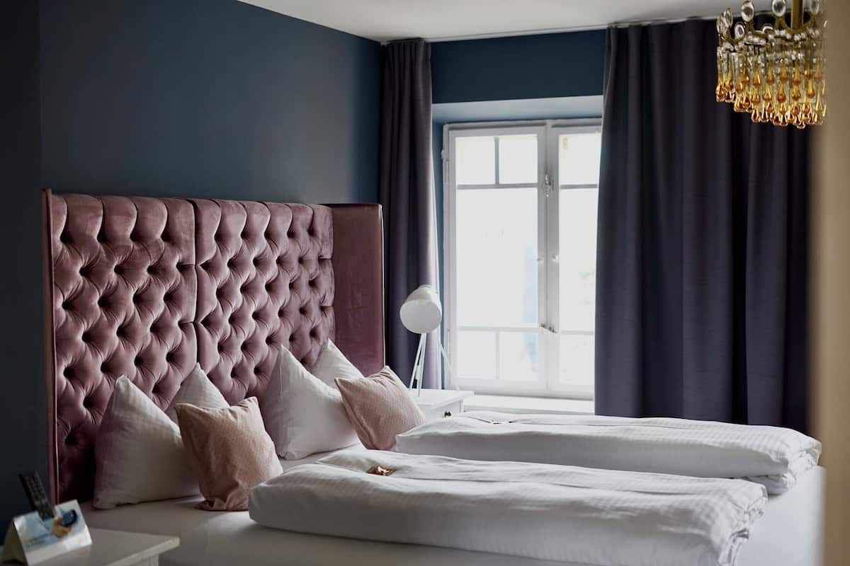 A bedroom with a tufted headboard, a large window with curtains, and a bedside lamp.