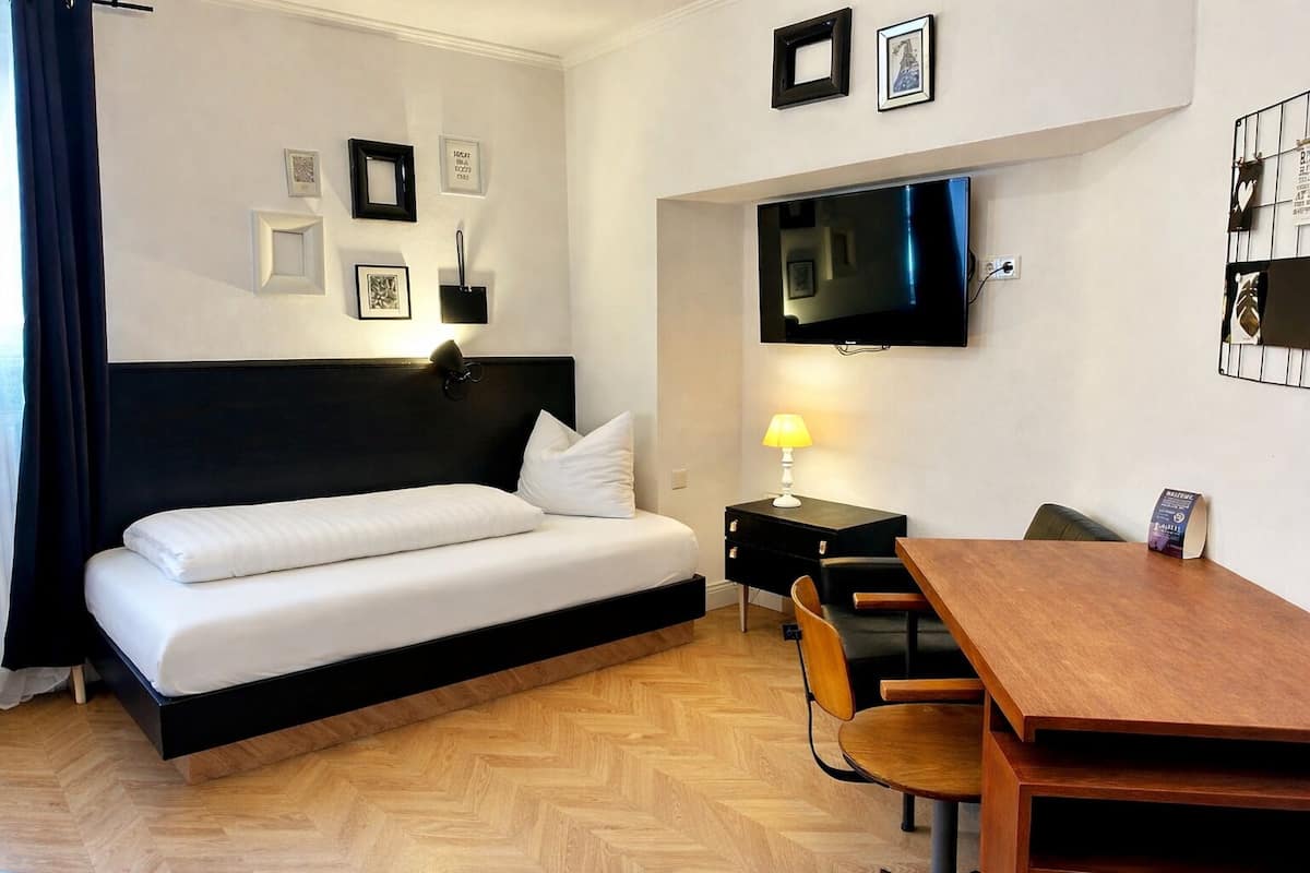 A hotel room with a bed, a desk, a chair, a TV mounted on the wall, and a wooden floor.