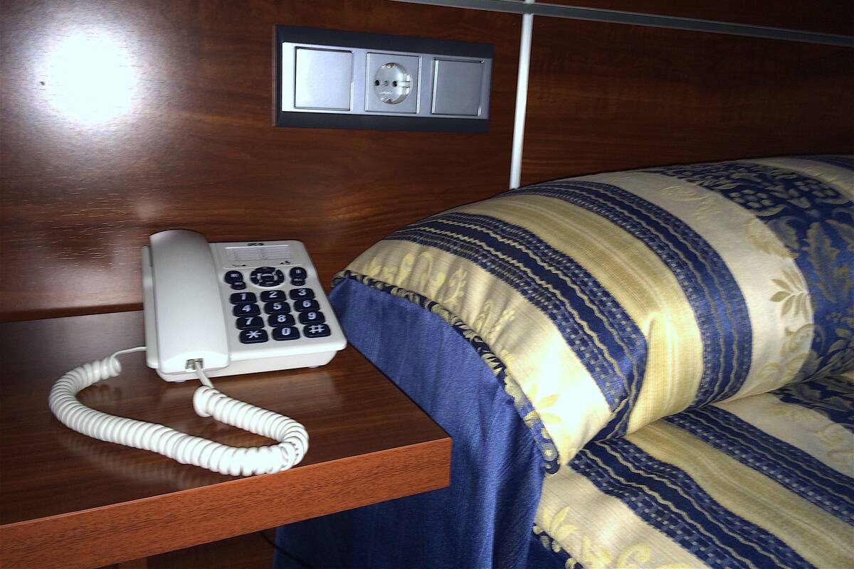 A hotel room with a wooden bedside table, a white telephone, and a bed with a blue and yellow striped blanket.