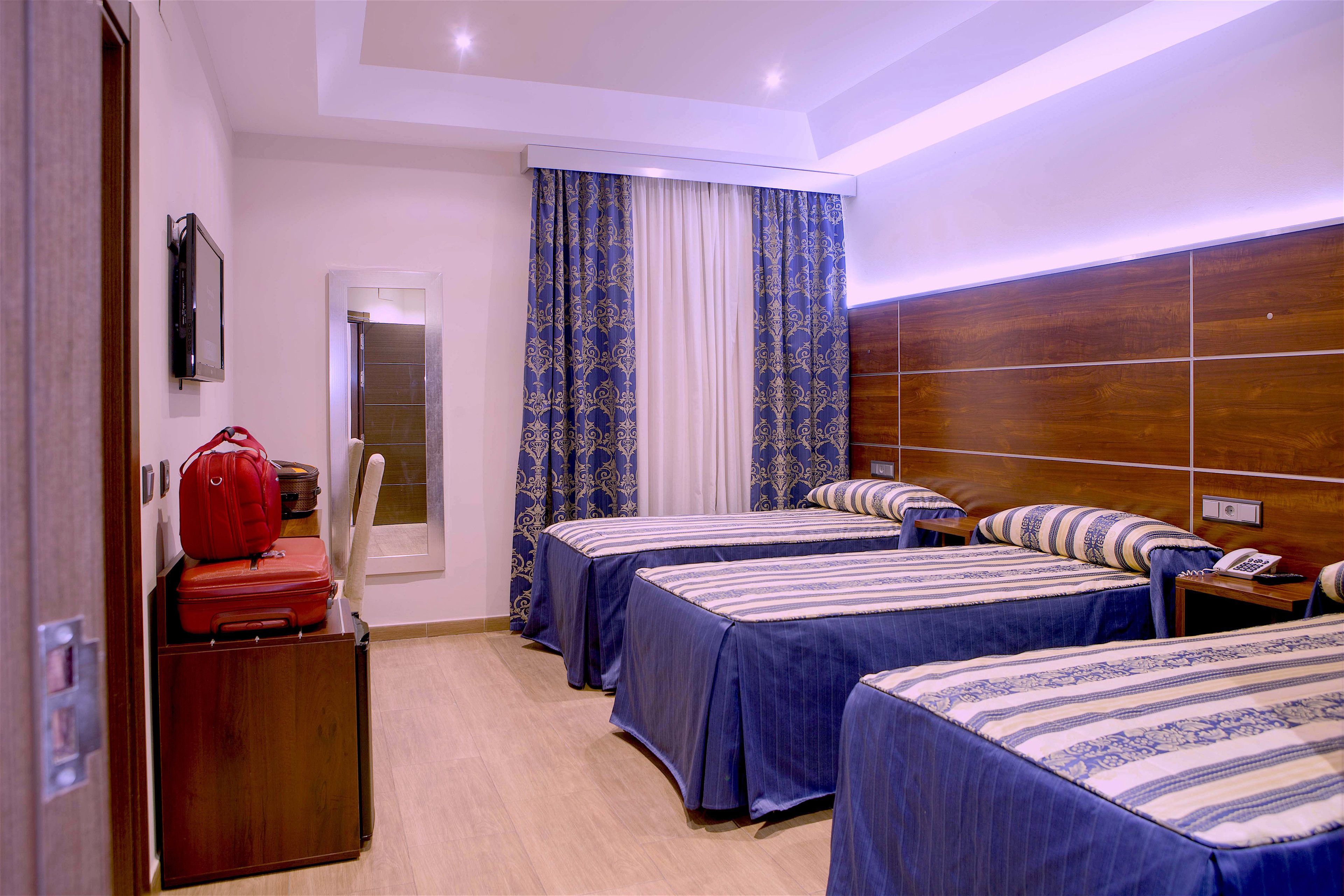 A hotel room with two beds, a wooden headboard, a red suitcase, and a TV on the wall.