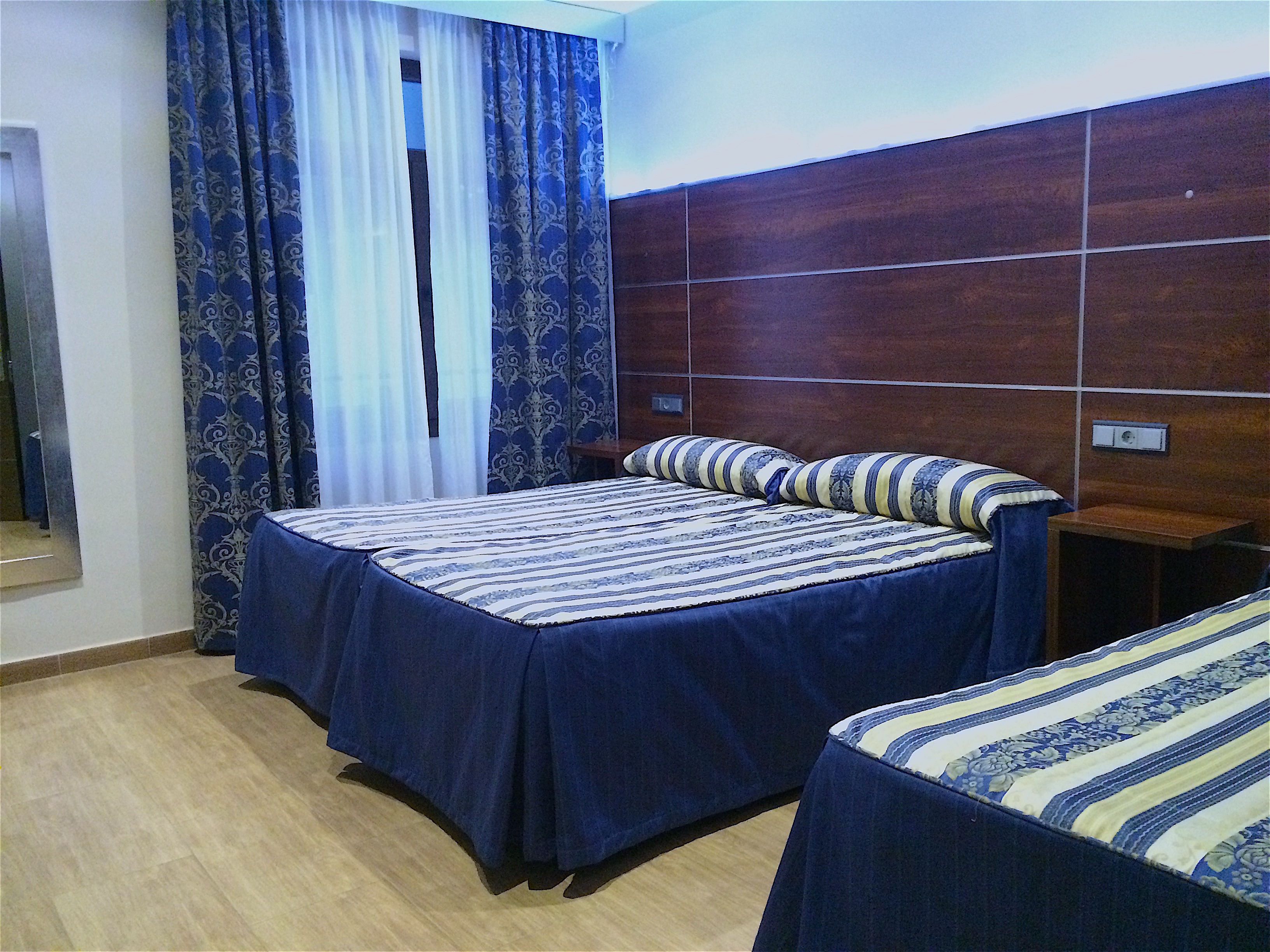 A hotel room with a double bed, a smaller bed, a wooden headboard, a nightstand, and a window with patterned curtains.
