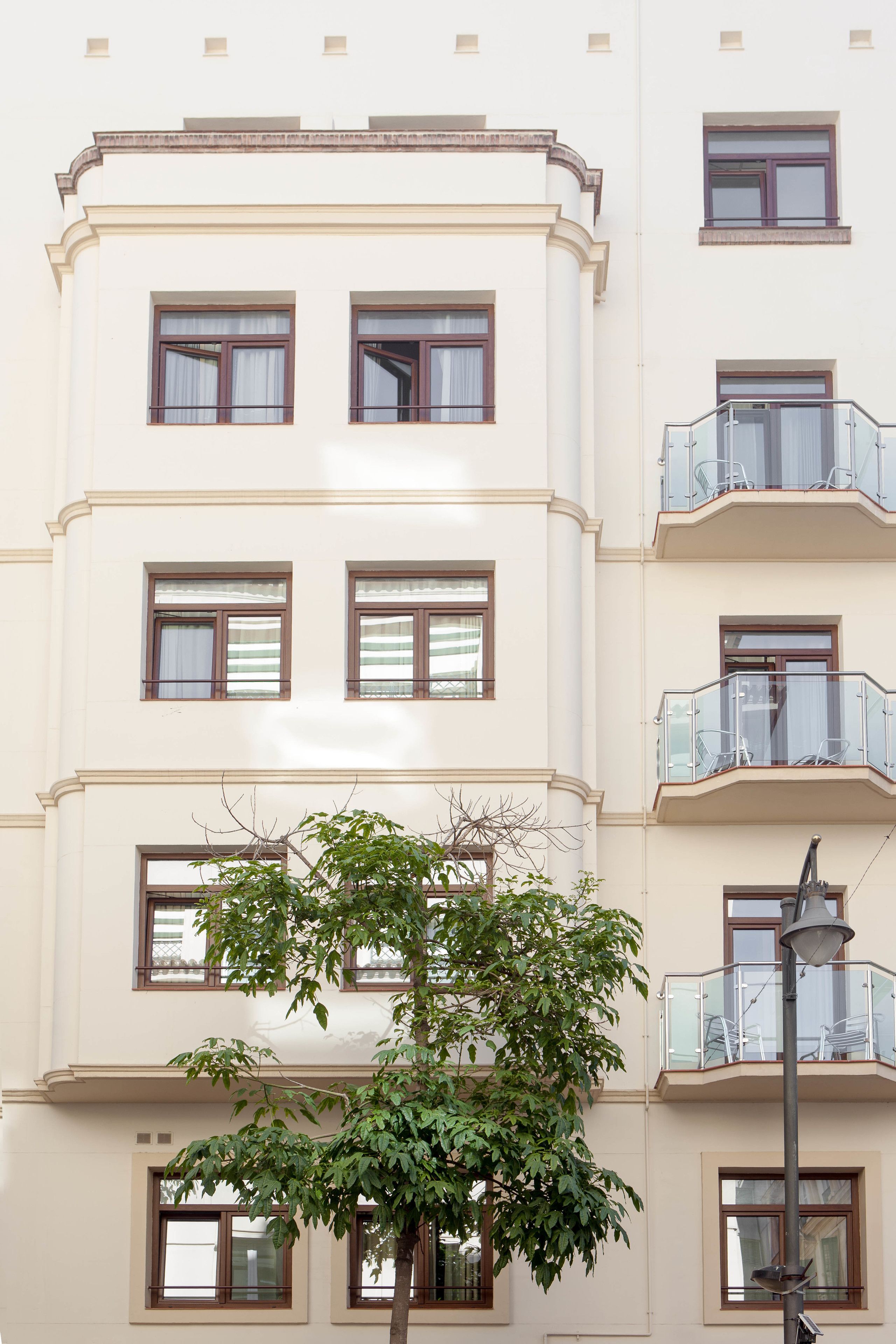 A multi-story building with balconies, large windows, and a tree in front.