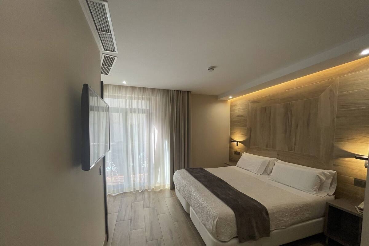 A modern hotel room with a large bed, a flat-screen TV mounted on the wall, and a wooden headboard.