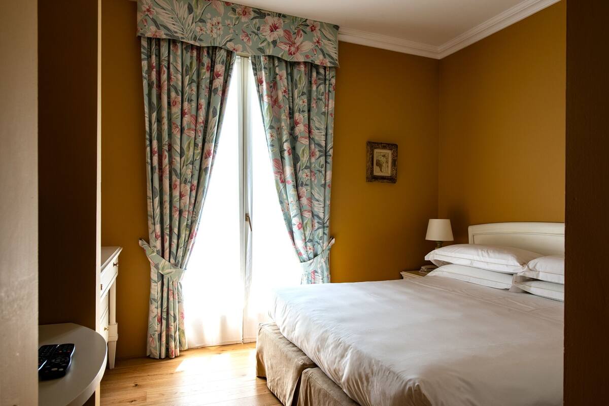 A bedroom with a bed, a bedside table, a lamp, and a window with floral curtains.