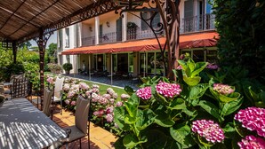 A covered outdoor seating area with tables and chairs, surrounded by lush greenery and pink flowers. - Hotel Franceschi (Forte dei Marmi)