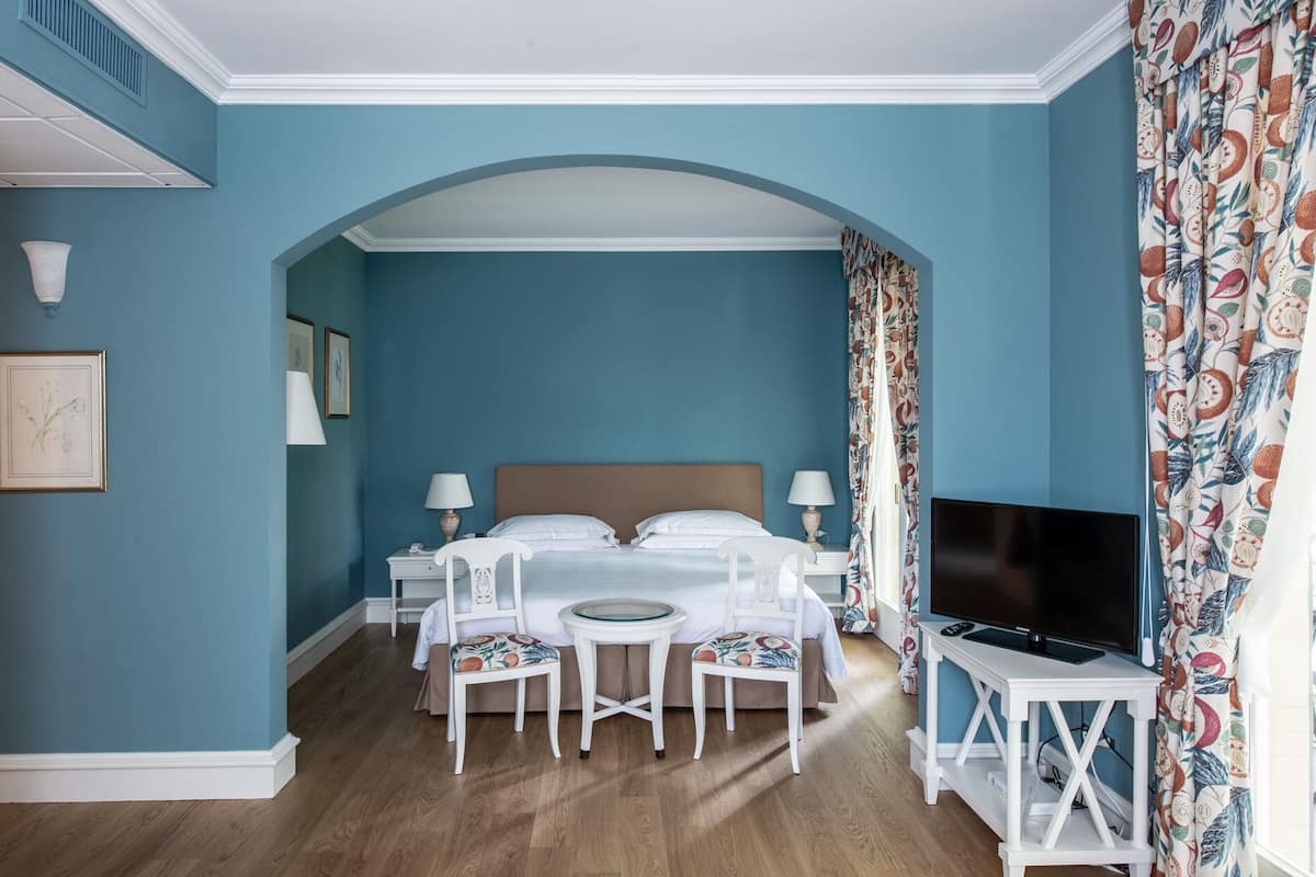 A hotel room with a bed, two bedside tables with lamps, a TV on a stand, a small round table with a chair, and a large window with curtains.