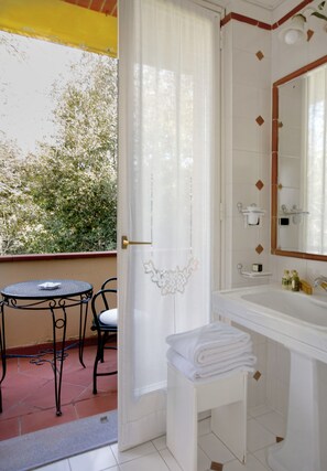 A bathroom with a white sink, mirror, and towel rack. There is a small table and chair outside the bathroom. - Hotel Franceschi (Forte dei Marmi)