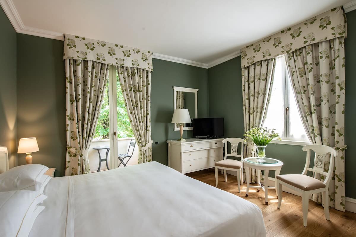 A hotel room with a bed, a desk with a TV, a chair, a small table with a vase, and curtains with a floral pattern.