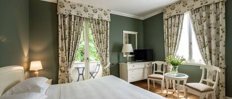 A hotel room with a bed, a desk with a TV, a chair, a small table with a vase, and curtains with a floral pattern.