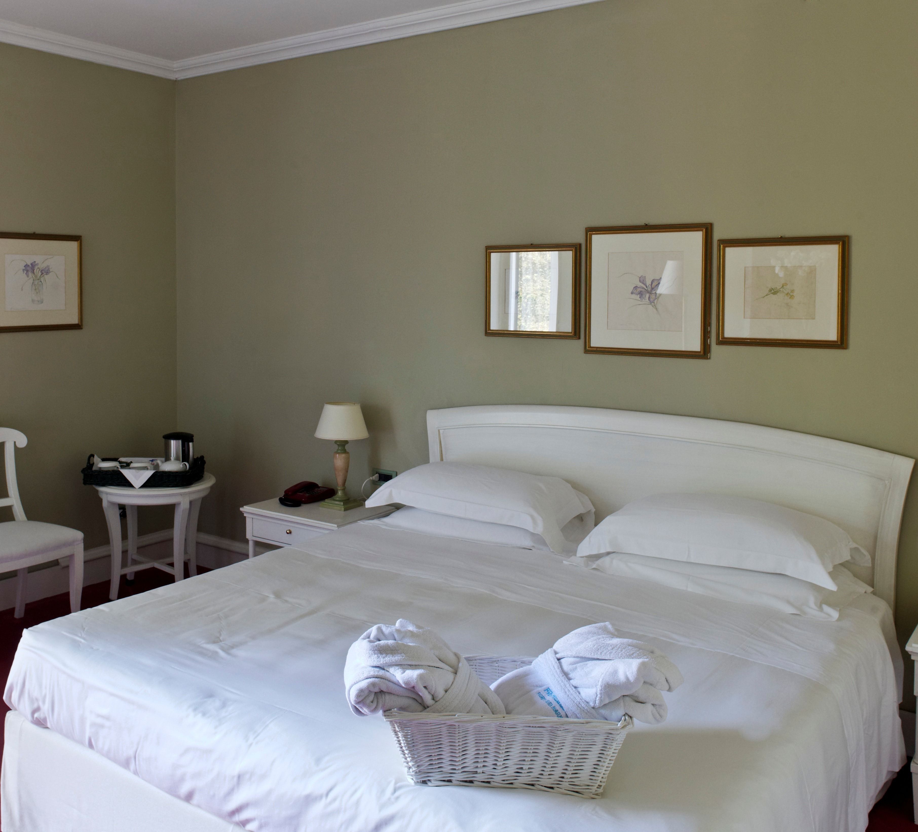 Una cama bien hecha con sábanas blancas, una mesita de noche con lámpara, una silla y una pequeña mesa con teléfono y cesta.