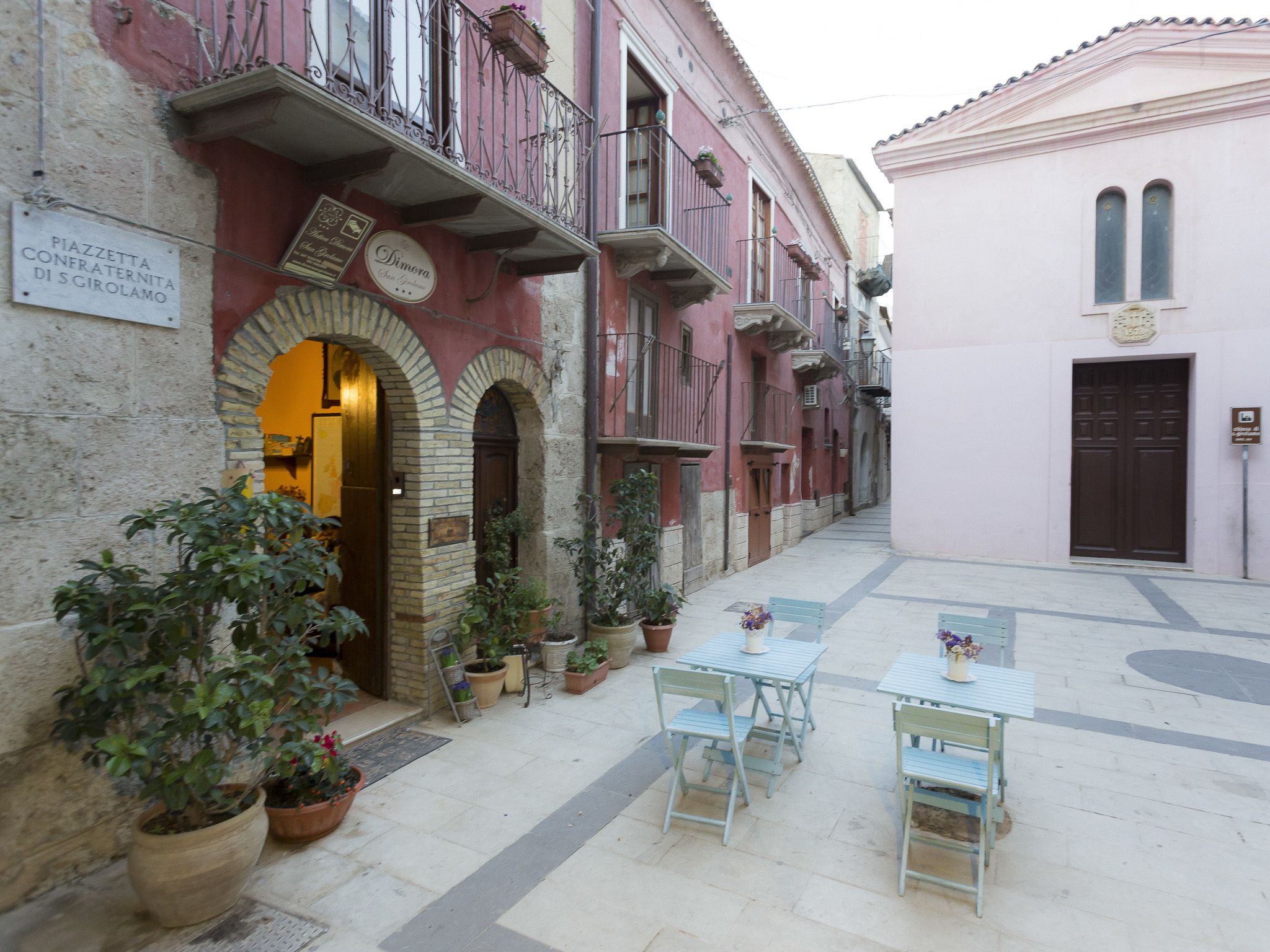 A narrow street with buildings on either side, outdoor seating, and potted plants.
