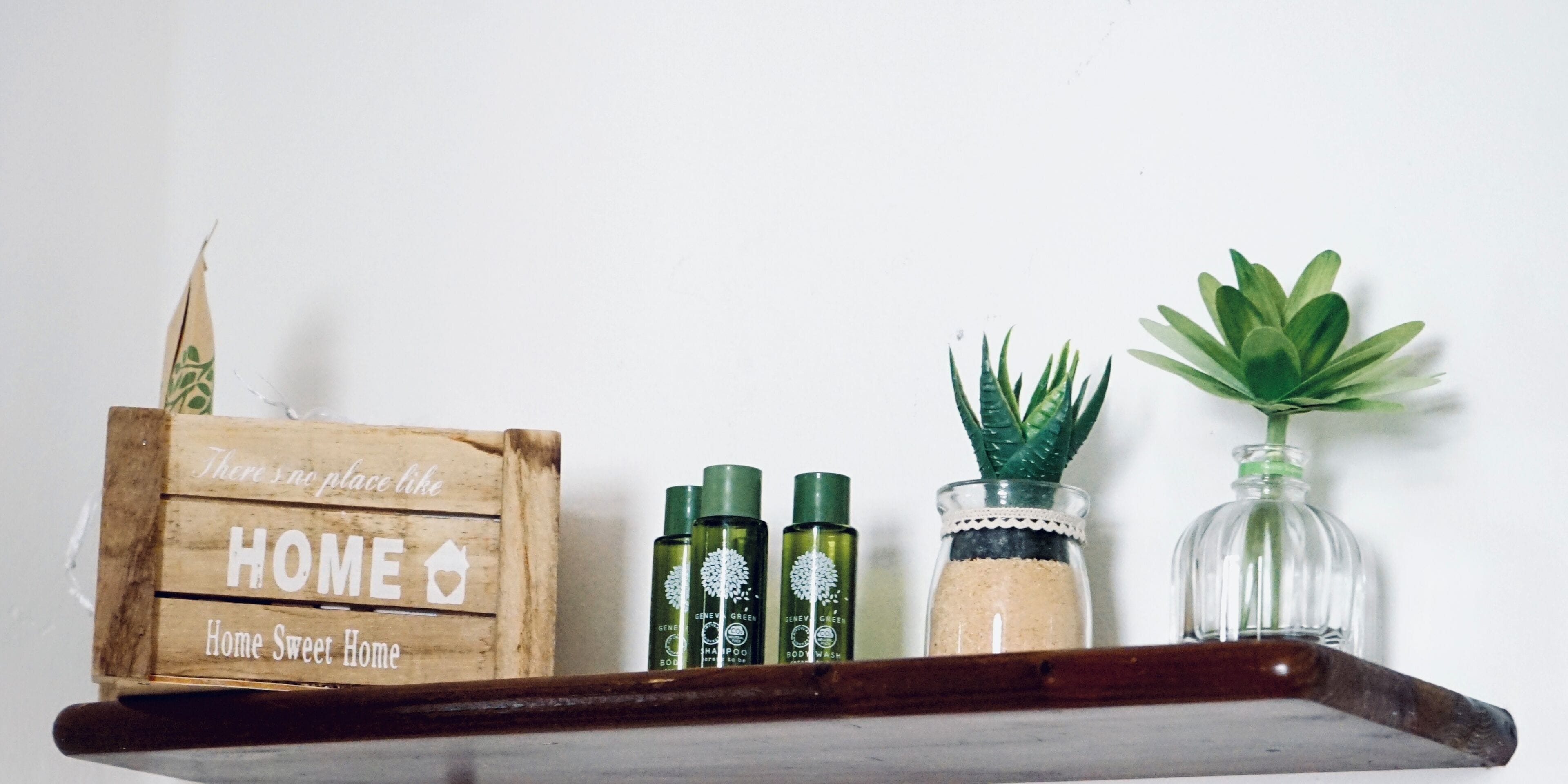 A wooden crate labeled "Home Sweet Home" with three bottles of green body wash, a glass jar with sand, and a small potted plant on a shelf.