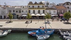 A waterfront area with boats docked, a café with outdoor seating, and a building with a yellow stripe. - Residencial Marés (Tavira)