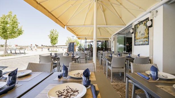An outdoor dining area with tables and chairs under a large umbrella, overlooking a beach. - Residencial Marés (Tavira)