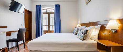 Habitación de hotel con cama, escritorio, silla, televisor, ventana con cortinas y lámpara.