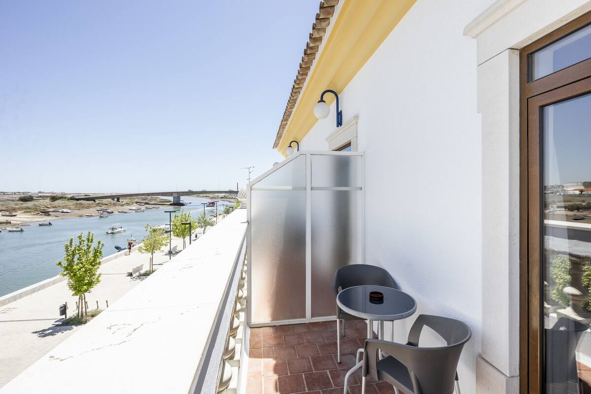 A balcony with a view of a river and boats, a small table, and chairs.