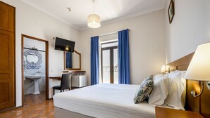 A hotel room with a bed, a desk, a chair, a TV, a bathroom with a sink and mirror, and a window with curtains. - Residencial Marés (Tavira)